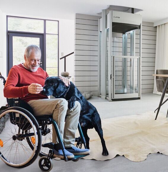 Mann im Rollstuhl füttert einen Hund aus der Hand, im Hintergrund ein Rollstuhllift